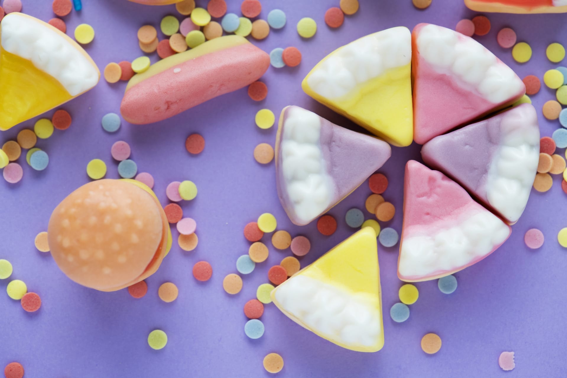 A cake made from sweets cut into 6 sections. The cake is sat on a background of sprinkled sweets and a burger made from jelly sweets.