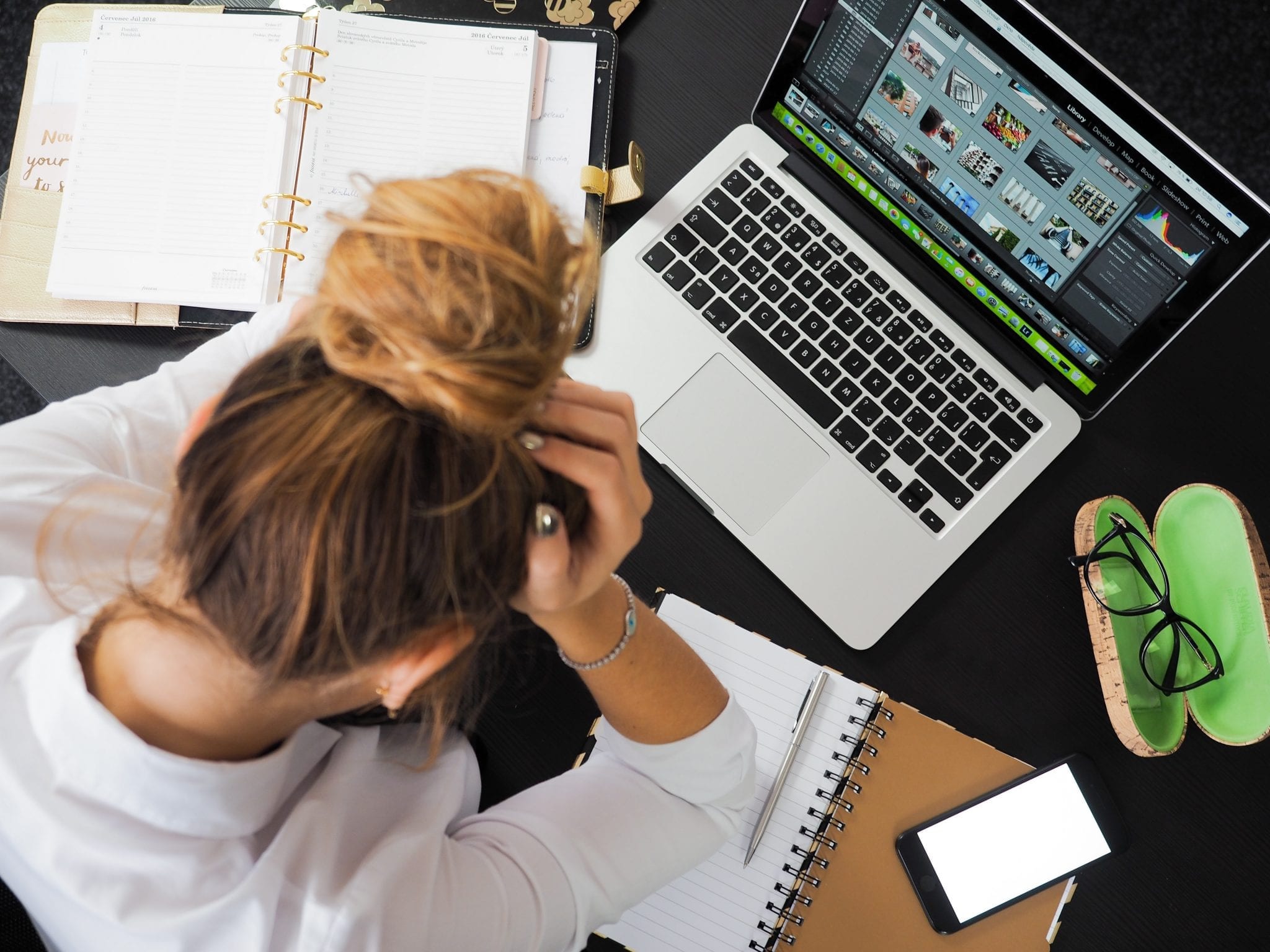 a lady sat at her desk with her head in her hands looking stressed. Her desk is cluttered with notebooks and a mac book