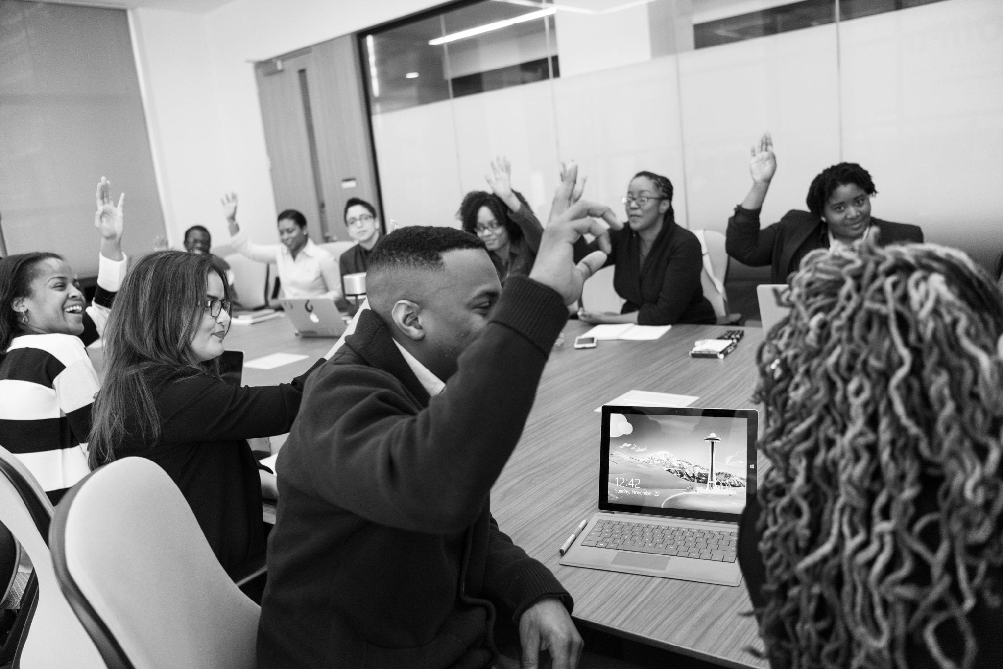 A board metting with 10 people all sat around a table. 7 of those people have their hands up.