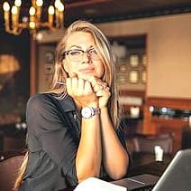 Blonde lady in glasses with hands to her chin looking thoughtful in front of a laptop
