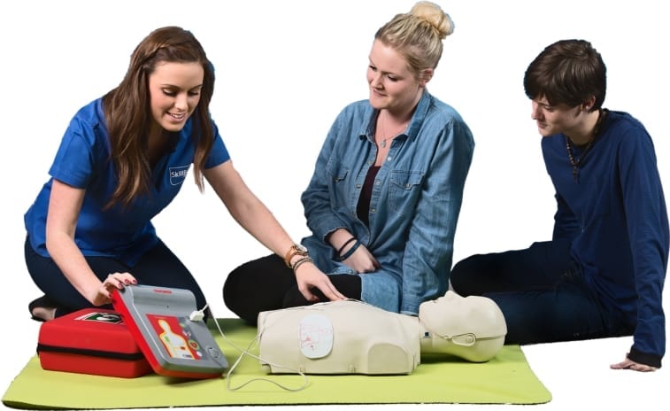 A lady in a skillbase first aid shirt showing two people how to use a defibrillator on a manikin