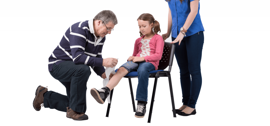 a small girl sits on a chair whilst a man bandages her right Knee. A woman is stood behind the chair overlooking the procedure.