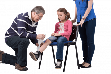 a small girl sits on a chair whilst a man bandages her right Knee. A woman is stood behind the chair overlooking the procedure.