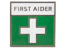 A small square pin badge that has a white cross on a green background and the text : First Aider