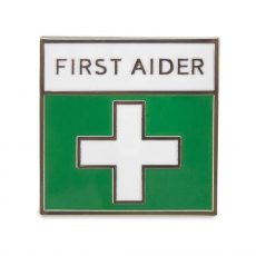 A small square pin badge that has a white cross on a green background and the text : First Aider