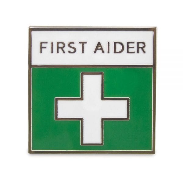 A small square pin badge that has a white cross on a green background and the text : First Aider