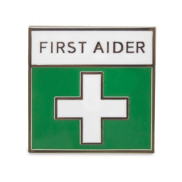 A small square pin badge that has a white cross on a green background and the text : First Aider