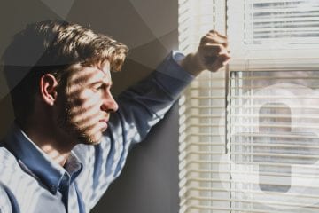 A man in a blue shirt looking out of a window whilst contemplating.