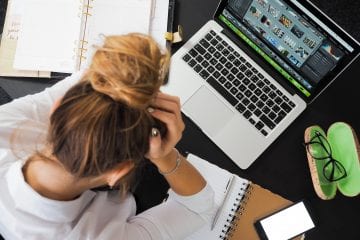 a lady sat at her desk with her head in her hands looking stressed. Her desk is cluttered with notebooks and a mac book