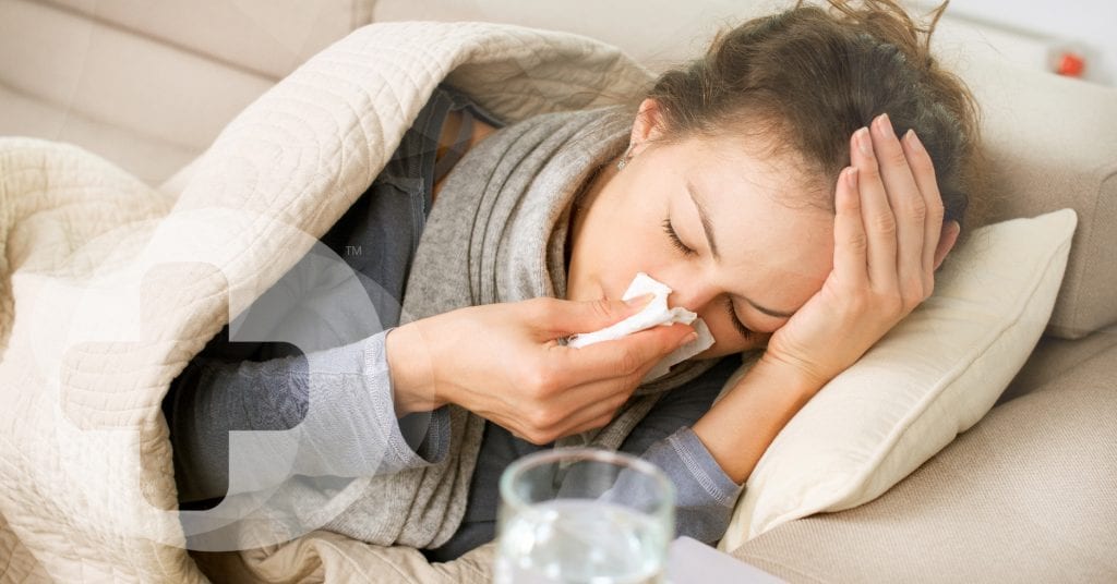 A lady who has the Coronavirus laying on a sofa under a blanket whilst blowing her nose.