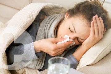 A lady who has the Coronavirus laying on a sofa under a blanket whilst blowing her nose.