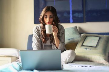 A woman in comfy clothes sat on a bed blowing into a cup whilst looking at a laptop.