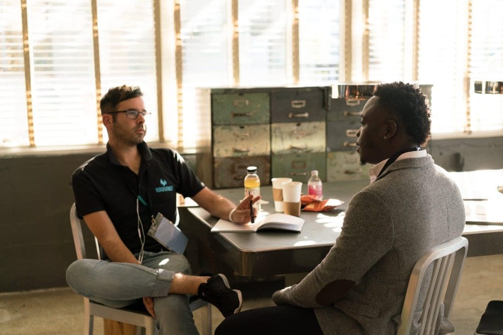 Two men sat at a table talking, one man has a note book open and a pen in his hand.