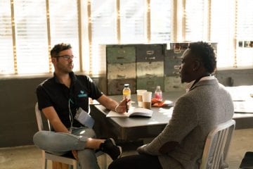 Two men sat at a table talking, one man has a note book open and a pen in his hand.